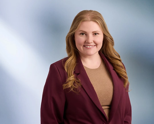“A woman, Mallory Reader, with shoulder-length, curly blonde hair, wearing a black blazer and white top, is smiling at the camera, standing against a blurred blue and white background.”