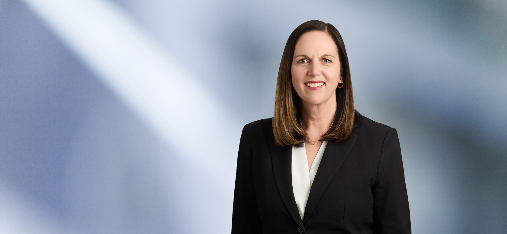 A woman, Anne Seurynck, in a black blazer and white blouse is smiling at the camera, standing against a blurred blue and white background.
