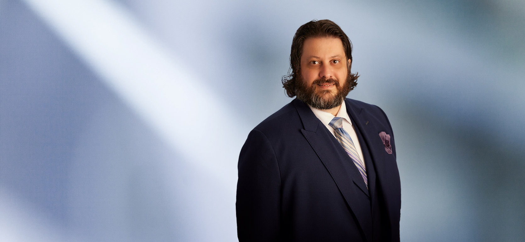 Smiling man, Robert Hamor, with medium-length dark hair and a beard, wearing a navy blue suit, striped tie, and pocket square, standing in front of a soft blue gradient background.