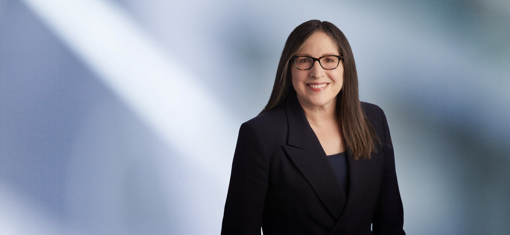 Smiling woman, Julie Fershtman, with long straight brown hair and glasses, wearing a dark blazer, standing in front of a soft blue gradient background