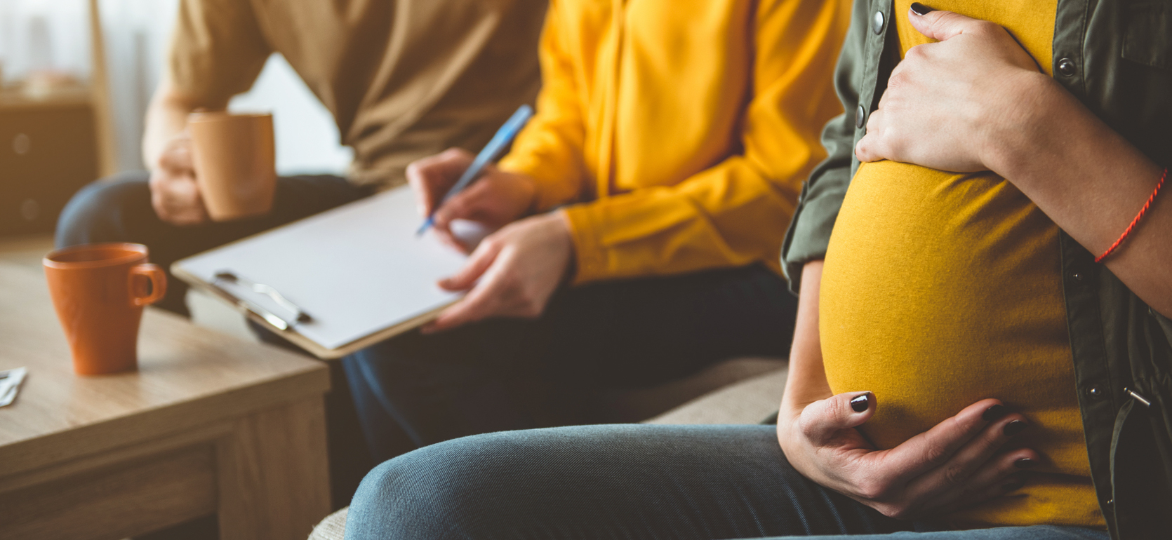 Three people sitting down with cups of coffee. One holds clipboard and pen and the other holds pregnant belly