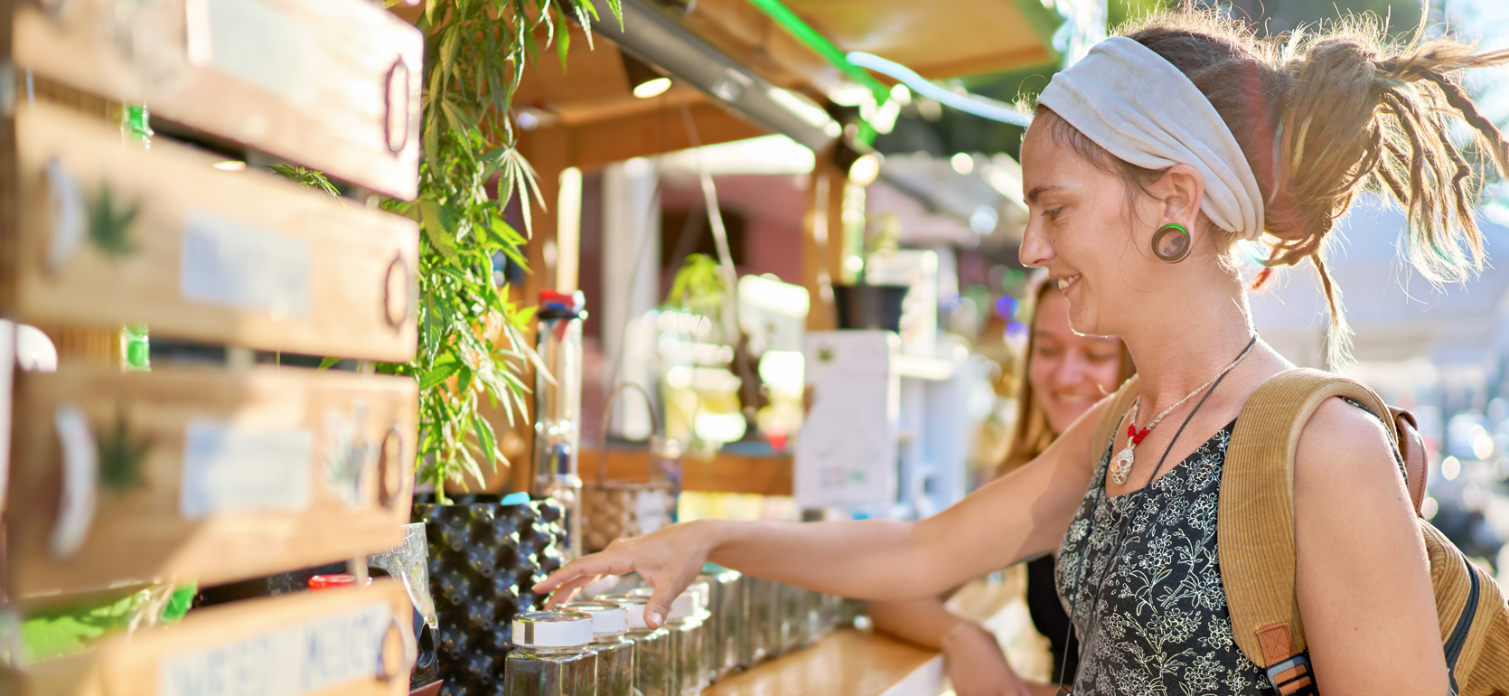 Young women with dreadlocks browse cannabis products at retail stand.