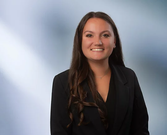 A young dark-haired woman, Rachel Conner, wearing a black sport coat and black blouse, smiles at camera in front of blurred blue-white background