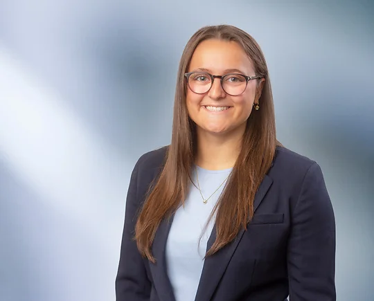 A young dark-haired woman with glasses, Olivia Reid, wearing a black sport coat and light blue blouse, smiles at camera in front of blurred blue-white background