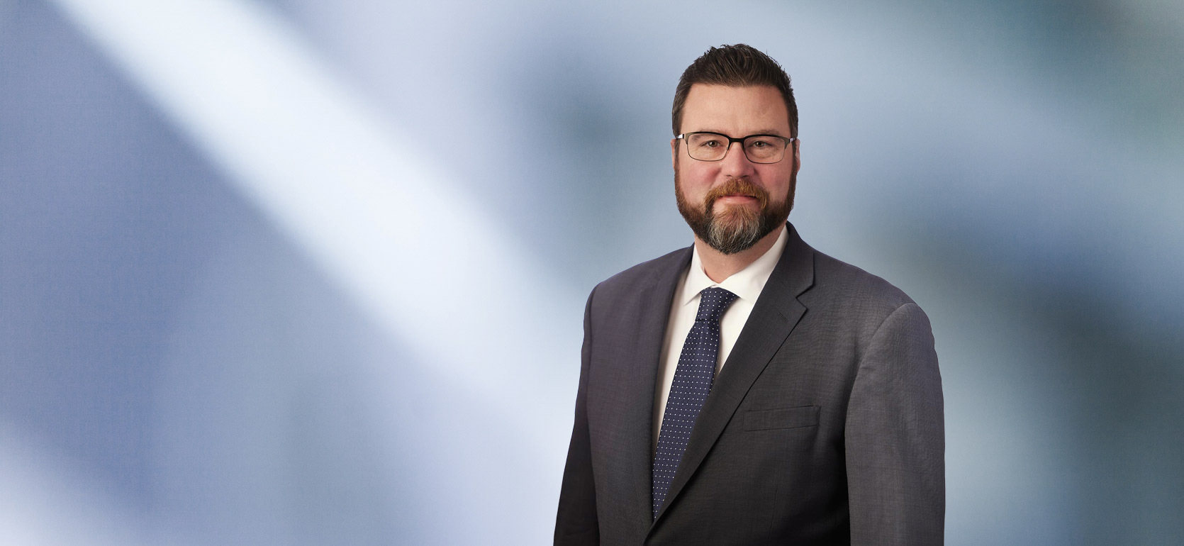“A man, Mark Koerner, with short, dark brown hair and glasses, wearing a grey blazer and white shirt, is smiling at the camera, standing against a blurred blue and white background.”