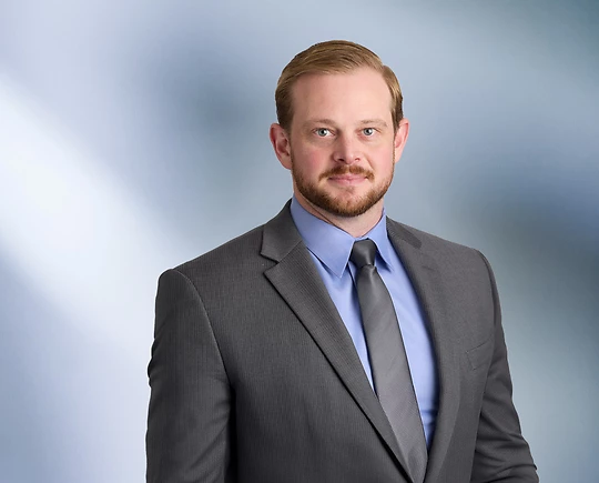A man, Michael Zahrt, in a dark suit, light shirt, and striped tie is smiling at the camera, standing against a blurred blue and white background.