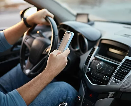A person driving a car is holding a smartphone in their right hand while steering with their left hand. The interior of the vehicle shows the dashboard, steering wheel, and center console. The person is wearing jeans and a long-sleeve shirt. This scene de