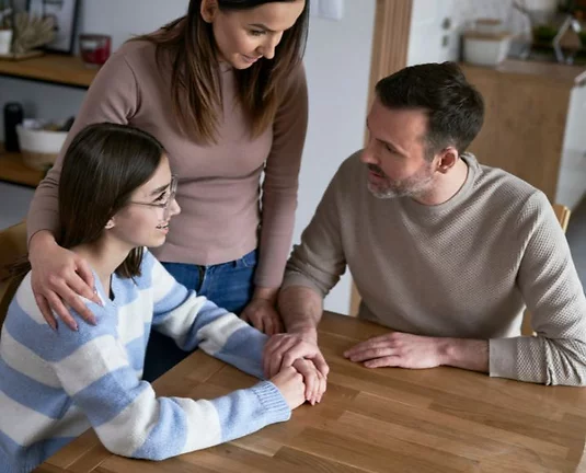 Parents sitting at a kitchen table having a warm, supportive conversation with their college-aged daughter, who is wearing glasses and a striped sweater. They are holding hands and smiling reassuringly.