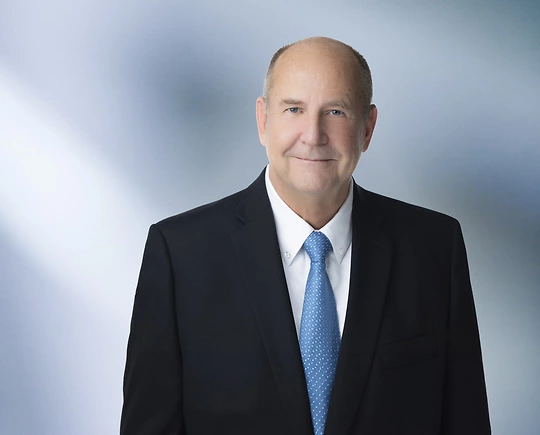A man, Bryan Cermak, in a dark suit, white shirt, and light blue tie is smiling at the camera, standing against a blurred blue and white background.