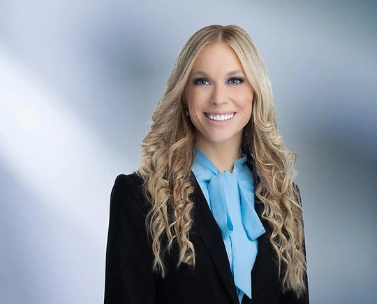 A woman, Jennifer Montasir, in a black blazer and bright blue blouse with long wavy blonde hair is smiling at the camera, standing against a blurred blue and white background.