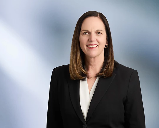A woman, Anne Seurynck, in a black blazer and white blouse is smiling at the camera, standing against a blurred blue and white background.