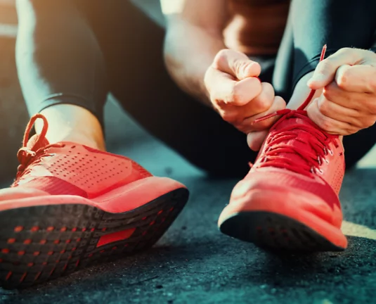 Close-up of a person tying the laces on bright red running shoes, sitting on pavement with sunlight casting a warm glow, preparing for physical activity or a run.
