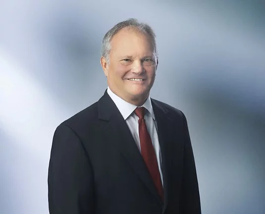 Professional portrait of a smiling middle-aged man, Brian Goodenough, in a dark suit, white shirt, and red tie, standing against a light gradient background.
