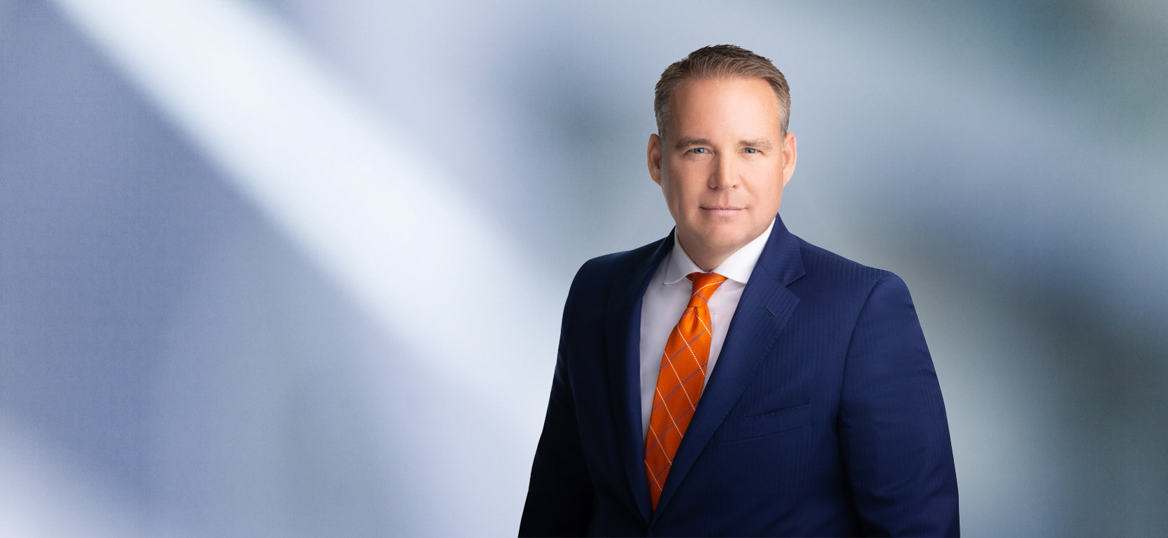 A man, Davis Russell, in a navy blue suit, white shirt, and orange patterned tie is looking at the camera, standing against a blurred blue and white background.