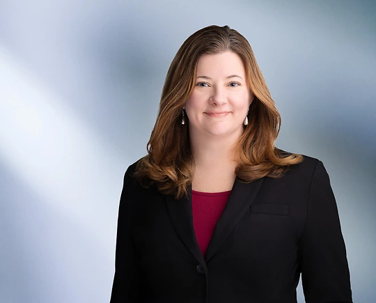 woman, Sara Cunningham, with light brown hair, wearing a black blazer, smiling against a blurred blue and white background"