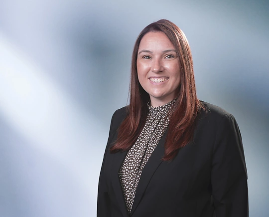 Woman, Stephanie Frizzell, wearing a dark blazer, standing against a blurred blue and white background.