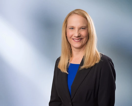 A woman, Lisa Myers, with long blonde hair, wearing a black blazer and bright blue top, is smiling at the camera, standing against a blurred blue and white background.