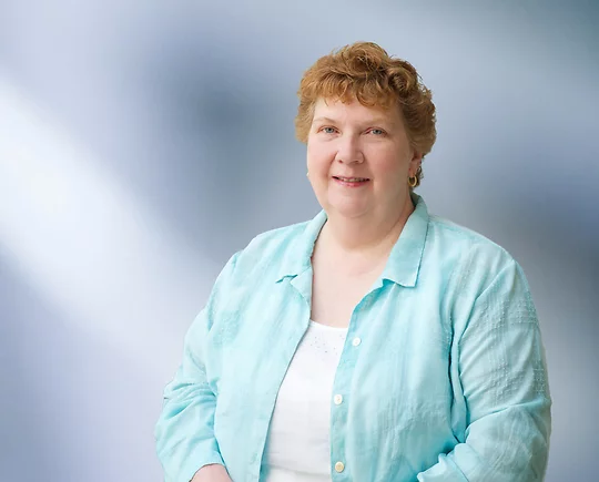 woman, Carole Schneider, wearing a light turquoise blouse, standing against a softly blurred blue and white background, smiling warmly at the camera.
