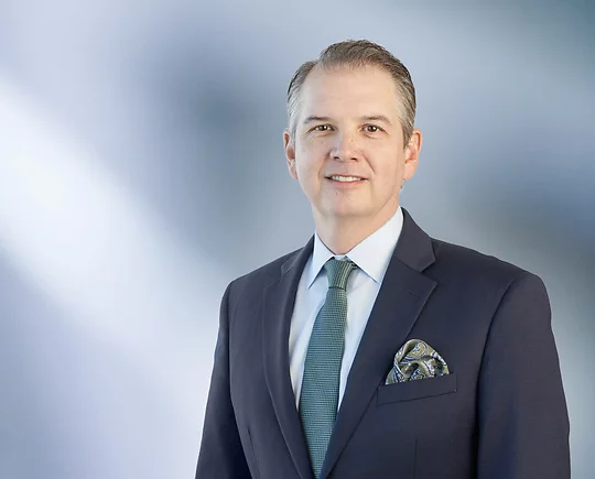 man, Matthew Fedor, wearing a dark suit, light blue shirt, green patterned tie, and matching pocket square, standing against a softly blurred blue and white background