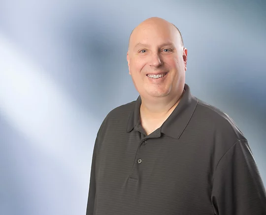Smiling bald man, John Bonandrini, wearing a dark grey polo shirt, standing in front of a soft blue gradient background.