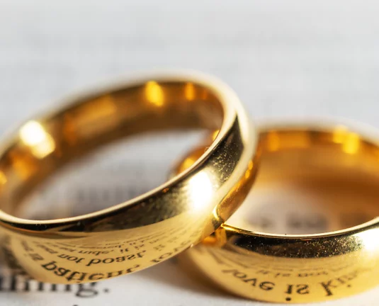 Two gold wedding rings resting on a document, symbolizing marriage or a legal union. One ring is slightly overlapping the other, and both have inscriptions on the inner bands.