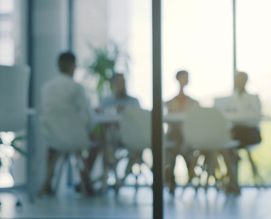 Blurry image of four people sitting around a conference table in a modern office with floor-to-ceiling windows, seen through glass walls.