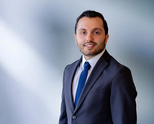 A young man, Badri Yono, wearing a blue-gray blazer, white shirt and solid blue time, smiles at camera in front of blurred blue-white background