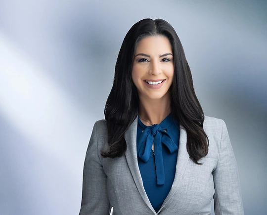 A woman, Courtney Agrusa, in a light gray blazer and blue blouse with a bow is smiling at the camera, standing against a blurred blue and white background.