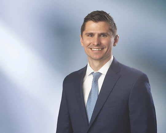 Man, Nicholas Oertel, with light brown hair wearing a charcoal blazer, smiling against a blurred white and blue background.