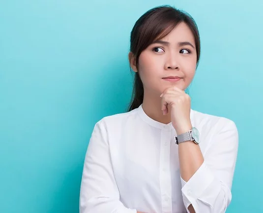 Thoughtful woman in a white blouse with her hand on her chin, looking to the side with a curious expression, standing against a light blue background.