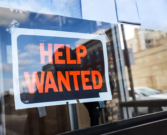 Black and white sign with orange letters that say Help Wanted taped to a window of a shop