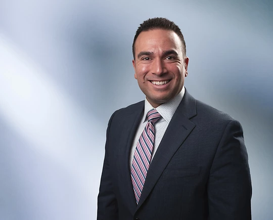 A man, Adam Fadly, in a dark suit, light shirt, and striped tie is smiling at the camera, standing against a blurred blue and white background.