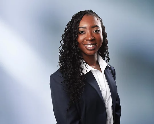 A woman, Kendra Dixie, in a dark navy blazer over a white button-up shirt is smiling at the camera, standing against a blurred blue and white background.