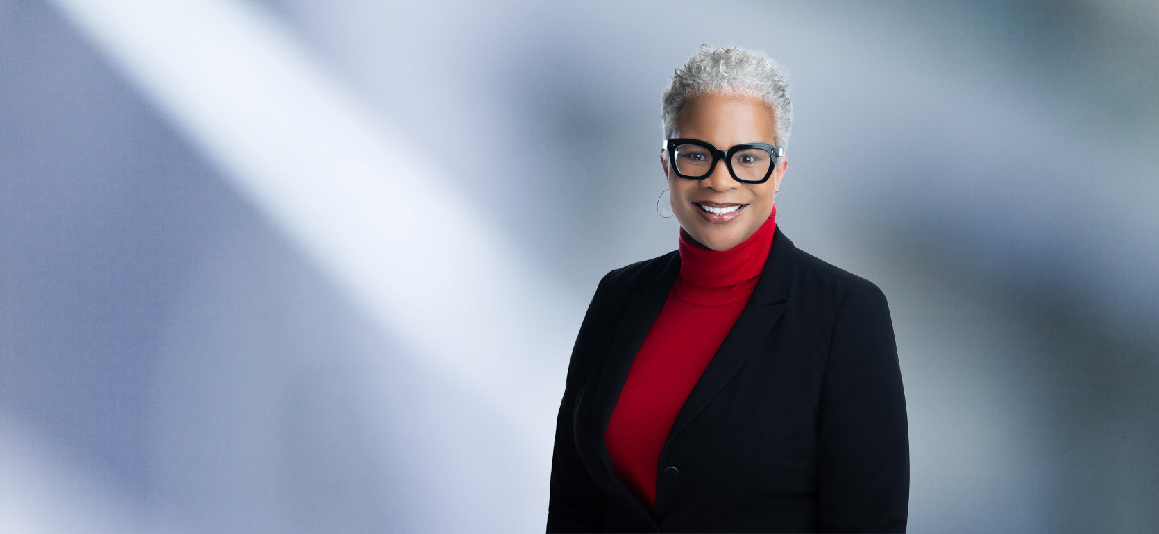 A woman, Dora Brantley, in a red turtleneck and black blazer is smiling at the camera, standing against a blurred blue and white background.