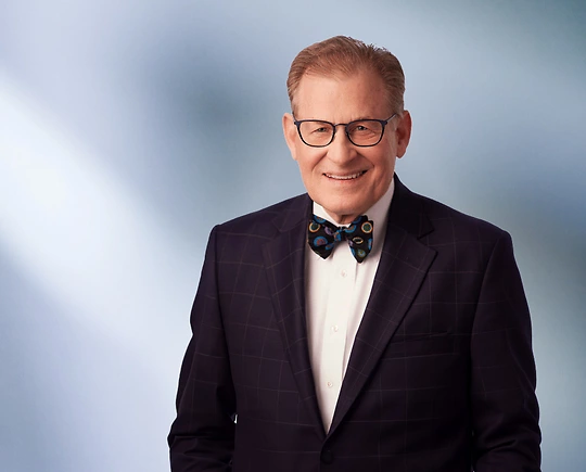 A man, Fredrick Dilley, in a dark checkered suit and a colorful bow tie is smiling at the camera, standing against a blurred blue and white background.