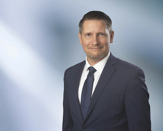 man, Warren Krueger, with short brown hair in a dark blazer, with a blurred blue and white background.