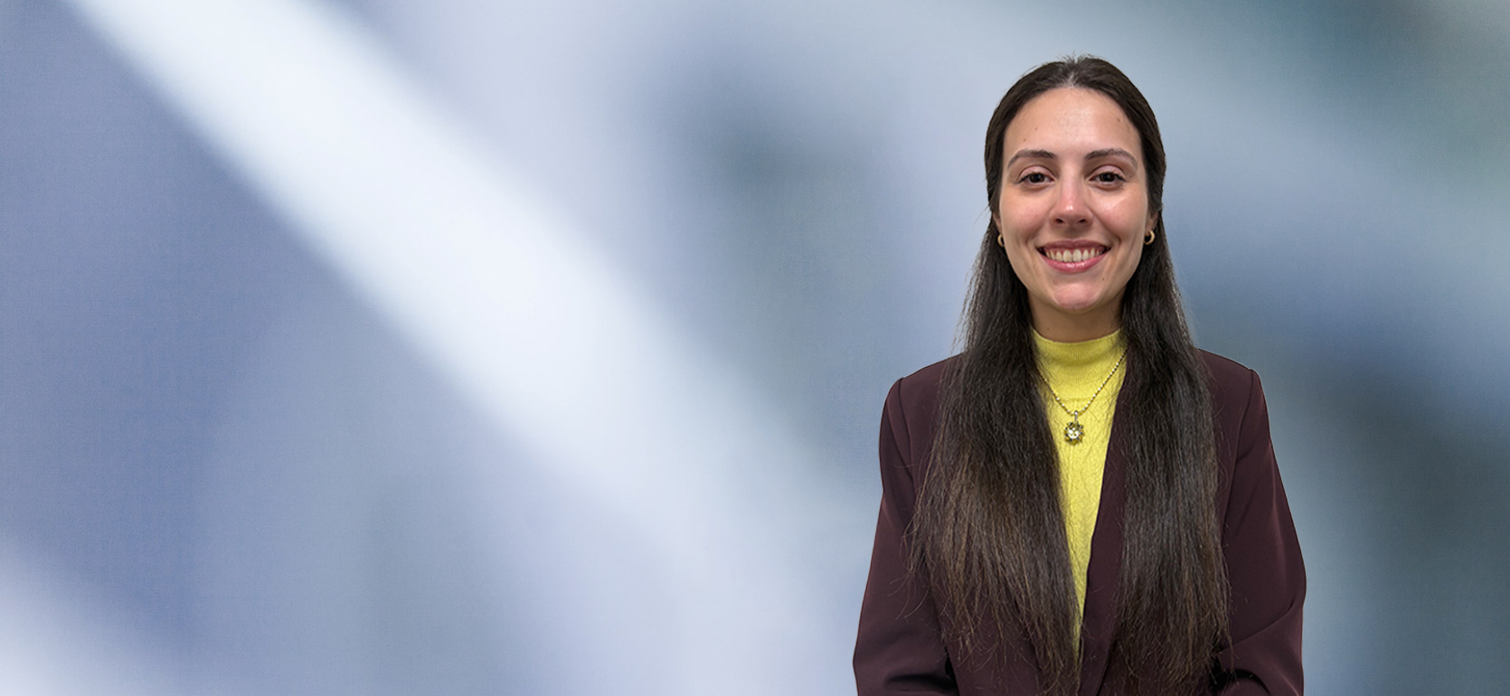 Woman with long brown hair, Stephanie Angelo, wearing a yellow turtleneck, necklace and purple blazer, smiles at camera in front of blurred blue gray background.