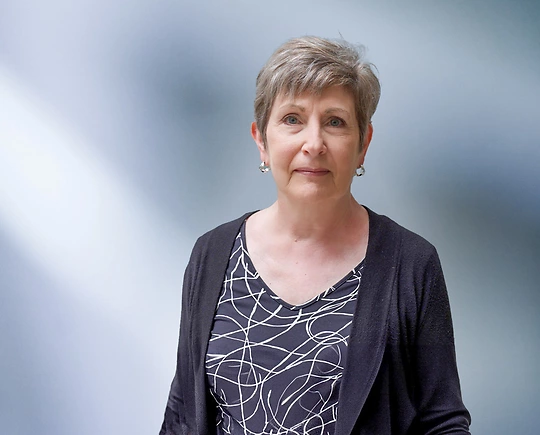 A woman, Carol Palomaki, in a black cardigan and patterned top is looking at the camera, standing against a blurred blue and white background.