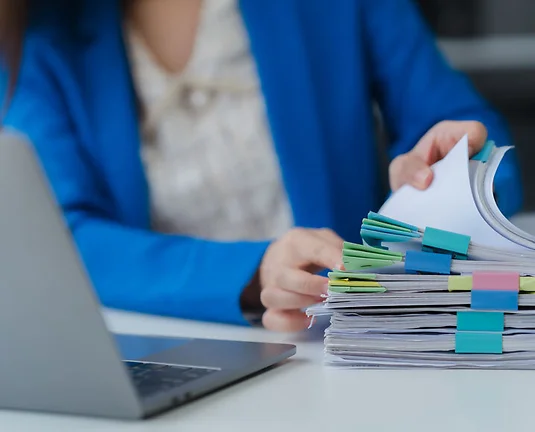 Workplace Documentation: A close-up shot of an office worker in a tailored blue blazer, meticulously reviewing a stack of documents.
