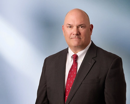 A man, Andrew Vredenburg, in a pinstripe suit, white shirt, and red patterned tie is looking at the camera, standing against a blurred blue and white background.