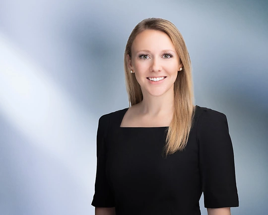 “A woman, McKenna Rievrs, with long, straight dark blonde hair, wearing a black top, is smiling at the camera, standing against a blurred blue and white background.”