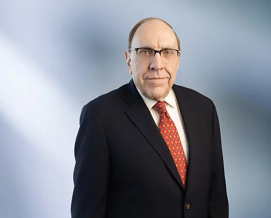 man, Brian Renaud, wearing a dark suit, white shirt, red patterned tie, and glasses, standing against a softly blurred blue and white background, looking confidently at the camera