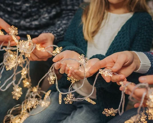 Close-up of multiple hands—children and adults—holding and untangling warm white holiday string lights shaped like snowflakes.