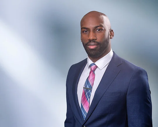 Bald bearded African American man, Xavier Prather, smiles at camera wearing a blue blazer, white undershirt and pink and blue striped necktie in front of blue gray blurred background.