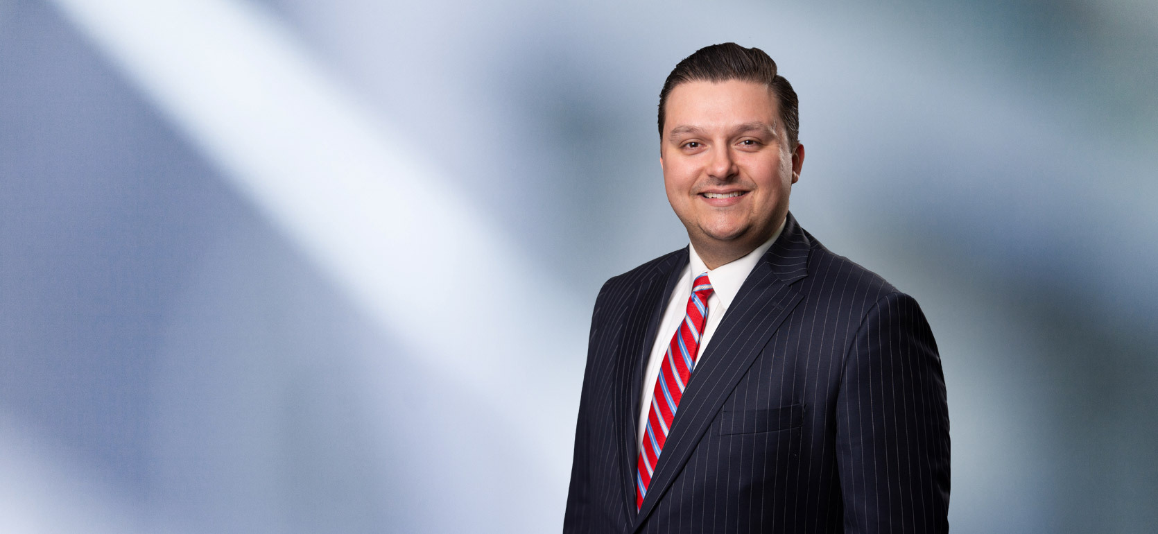 A man, Joe Giacalone, smiles at camera wearing a black pinstripe suit, white shirt and red striped tie in front of blue gray blurred background