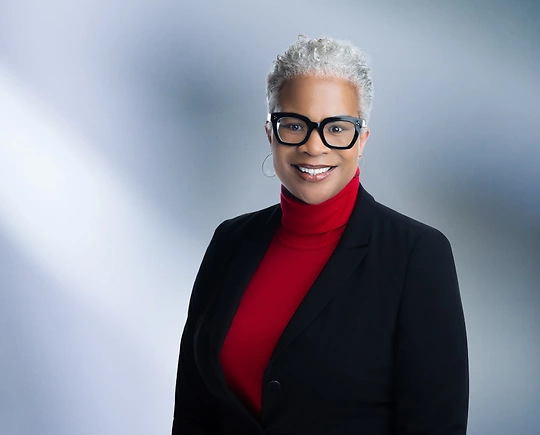 A woman, Dora Brantley, in a red turtleneck and black blazer is smiling at the camera, standing against a blurred blue and white background.