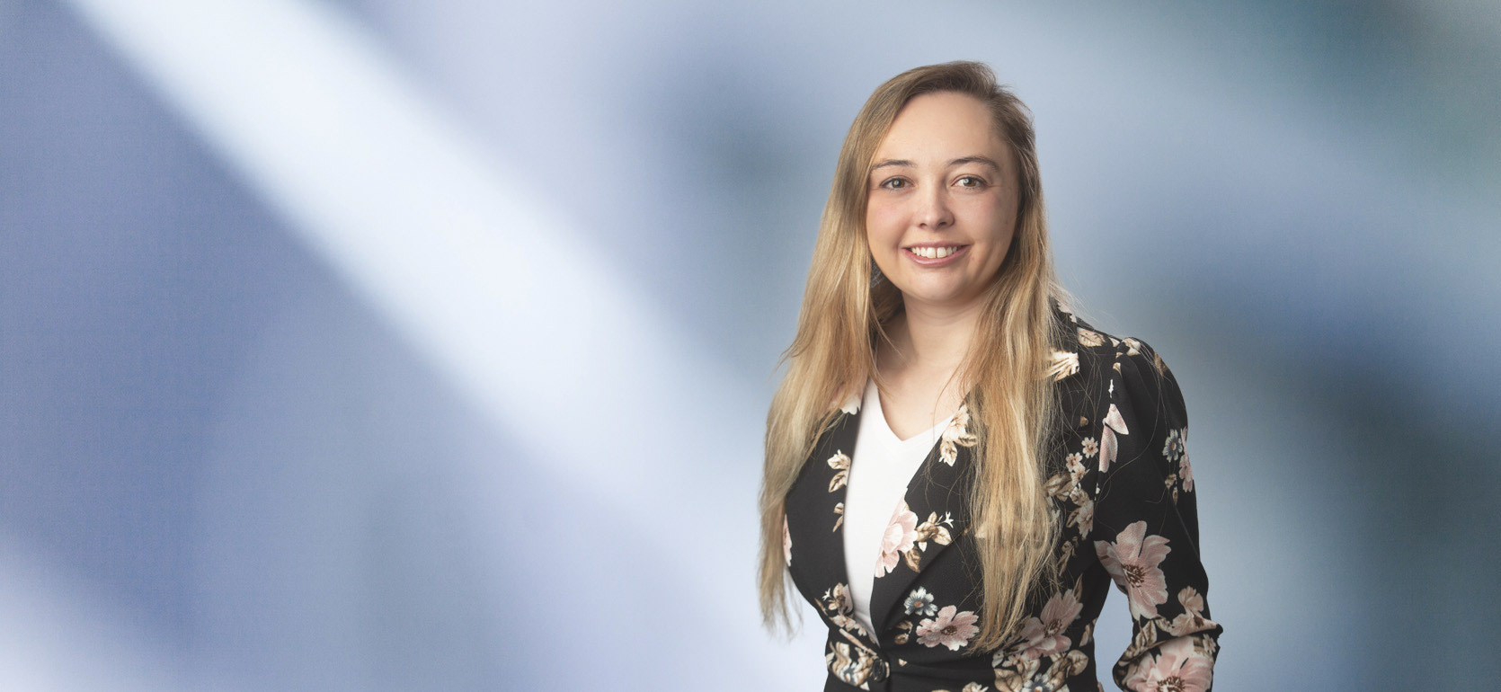 A woman, Jordan Asbury, in a black blazer with a white blouse and a floral pattern is smiling at the camera, standing against a blurred blue and white background.