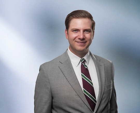 “A man, Matthew Murray, with short, light brown hair, wearing a grey blazer and white shirt, is smiling at the camera, standing against a blurred blue and white background.”