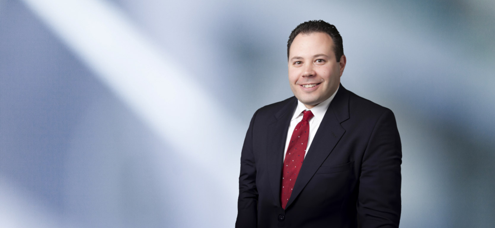 Smiling man, Ben Price, wearing a dark suit with a white shirt and red patterned tie, standing in front of a soft blue and white blurred background