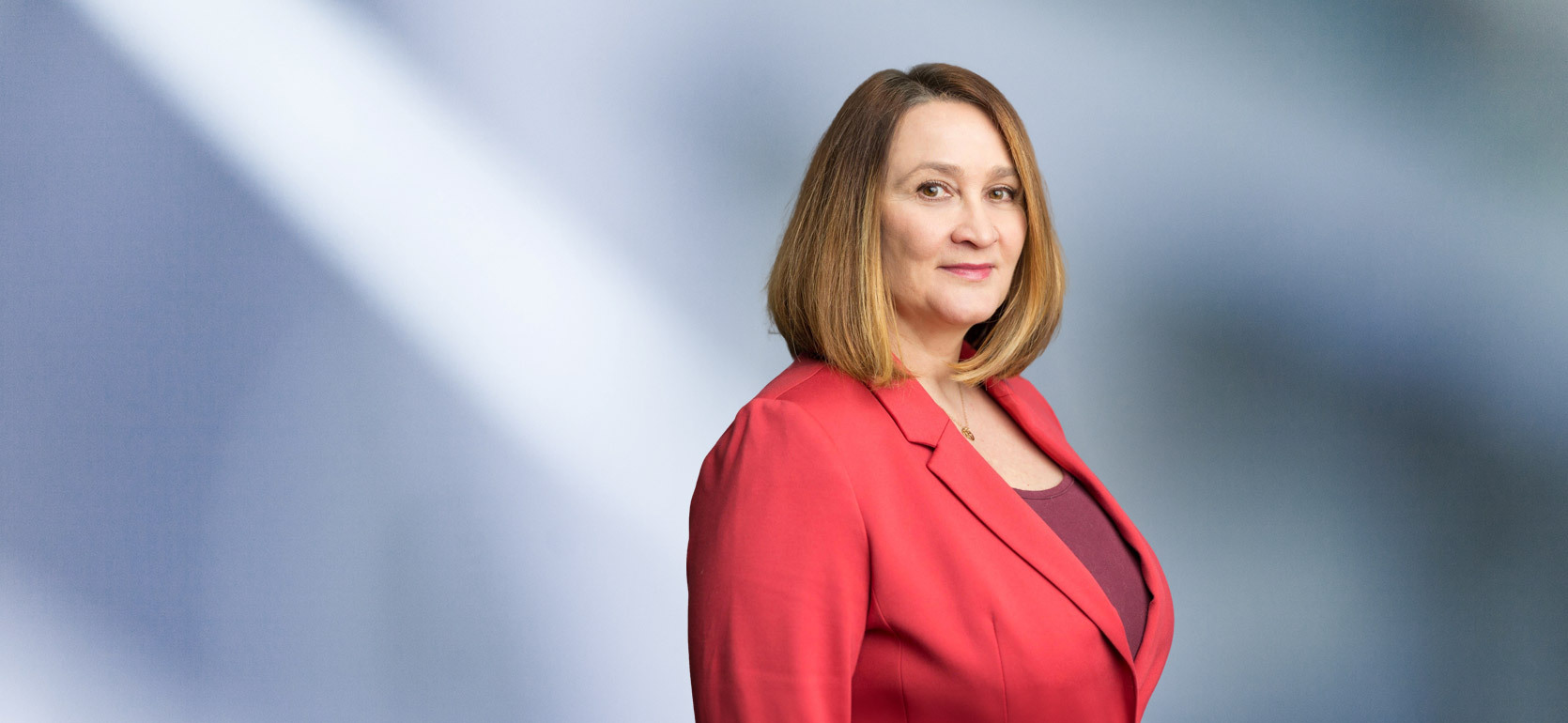 A woman, Inna Musser, in a red blazer and maroon top is looking at the camera, standing against a blurred blue and white background.